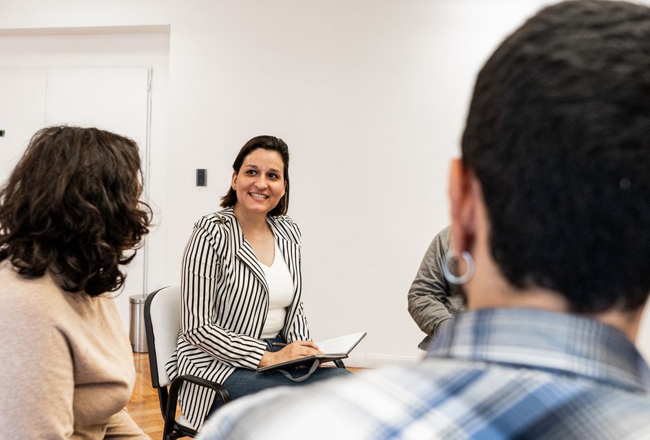 A woman with a notebook smiling as she talks to a group of people sat in a circle.