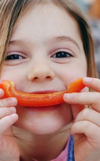 A young girl smiles while holding up a piece of red pepper up to her mouth. The piece of pepper recreates a smile.