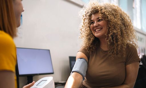 A woman with curly hair smiles while another woman checks her blood pressure in an office.