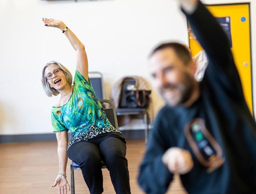 An elderly woman is sat down performing a stretch with her arms over her head.