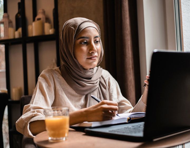 A young muslim woman wearing a hijab studies on a laptop while making notes in a notebook.