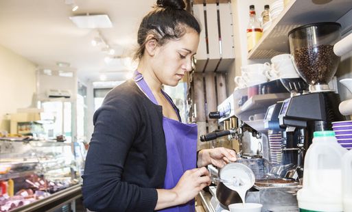 A barista in a cafe pouring steamed milk from a jug into a coffee cup