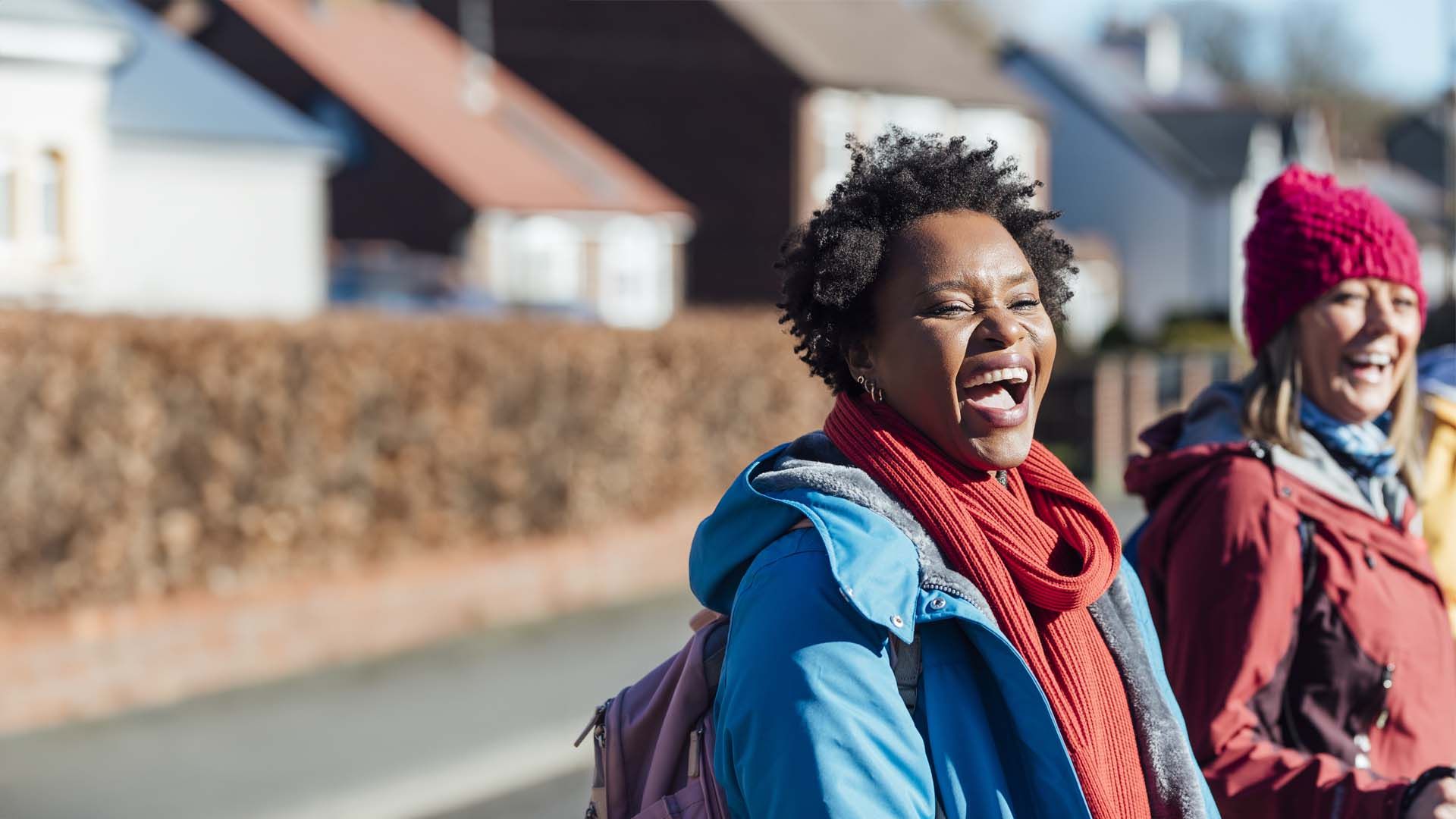A black woman with short curly black hair laughing. Two other women are blurred in the background.