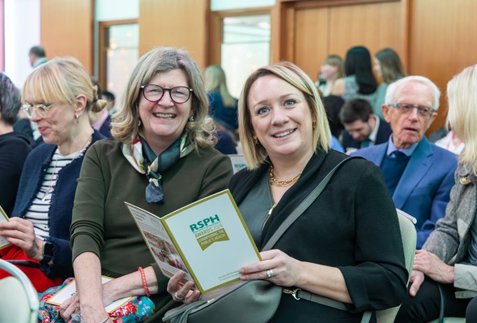 Two women sitting down at a busy Health & Wellbeing  Awards ceremony smiling to camera with one holding an event programme.