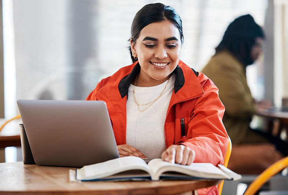 A woman wearing a red jacket uses a laptop with an open book next to her