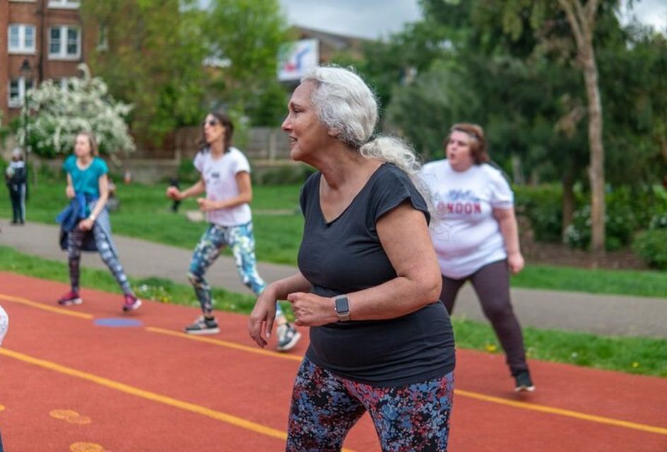An elderly white woman exercises in a park with others