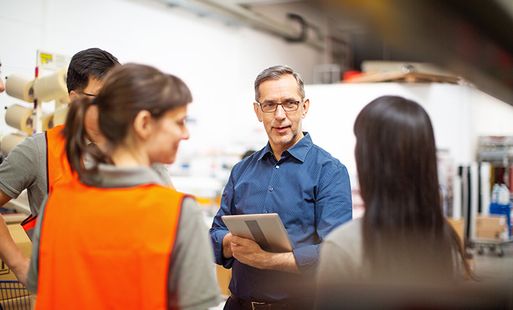 A man holding a tablet speaks to employees in a warehouse.