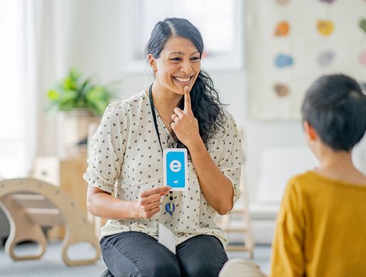 A speech therapist points at her mouth while holding up a card with the letter E on it.