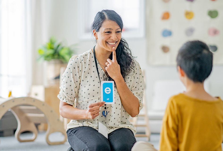 A speech therapist points at her mouth while holding up a card with the letter E on it.